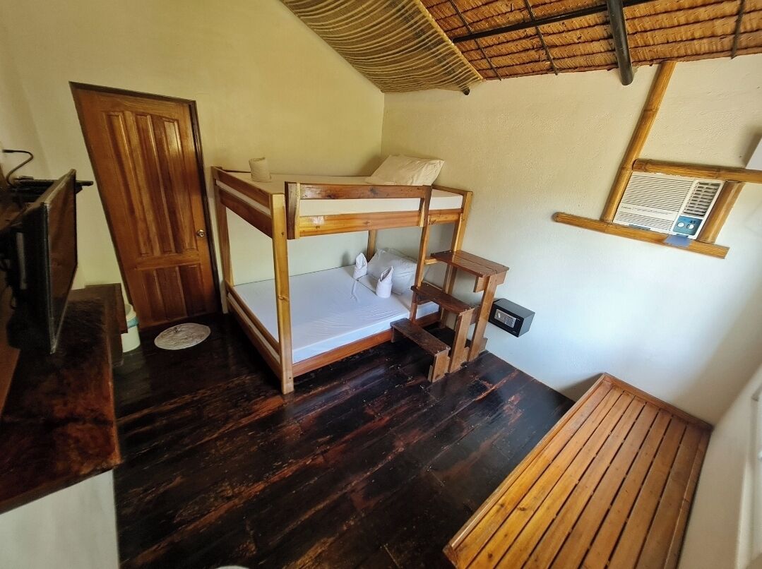 Een kleine, compacte kamer met een stapelbed, een houten bankje, een tafeltje, een deur en een raam met ventilatiesysteem.