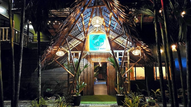 Una vista notturna dell'ingresso di un resort, con una struttura a tetto di paglia, un cartello con la scritta "Nativo D'Kubo" e un vialetto che conduce all'ingresso.