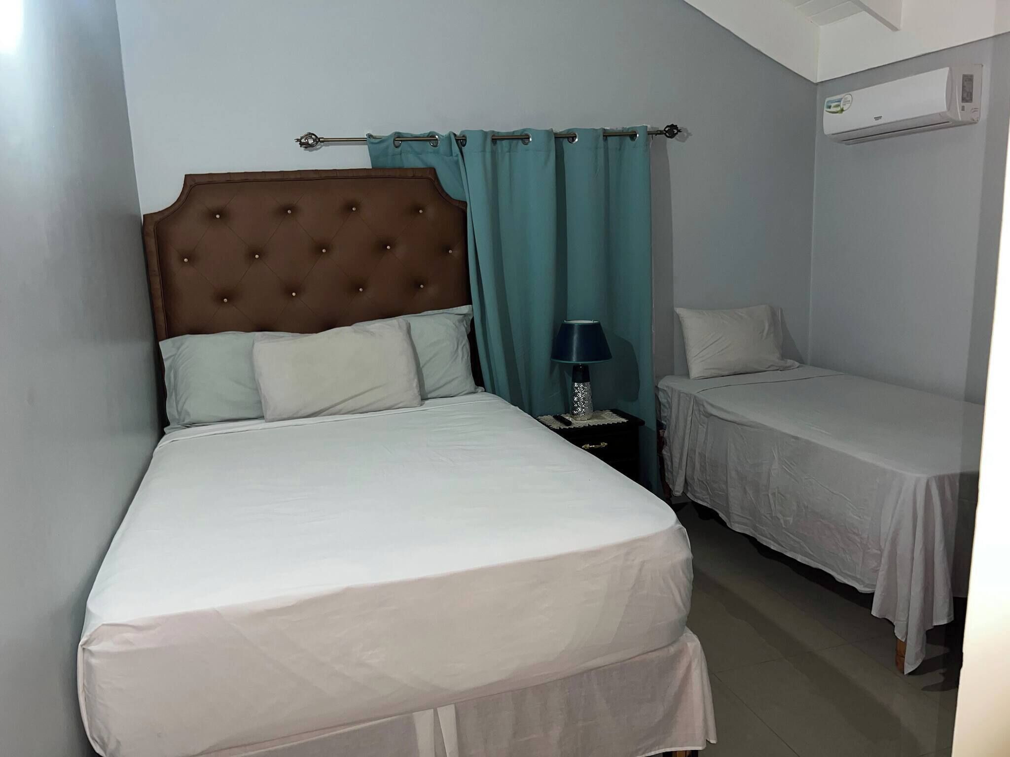 Une chambre avec un grand lit, un plus petit lit, une table de chevet avec une lampe et un appareil de climatisation fixé au mur.