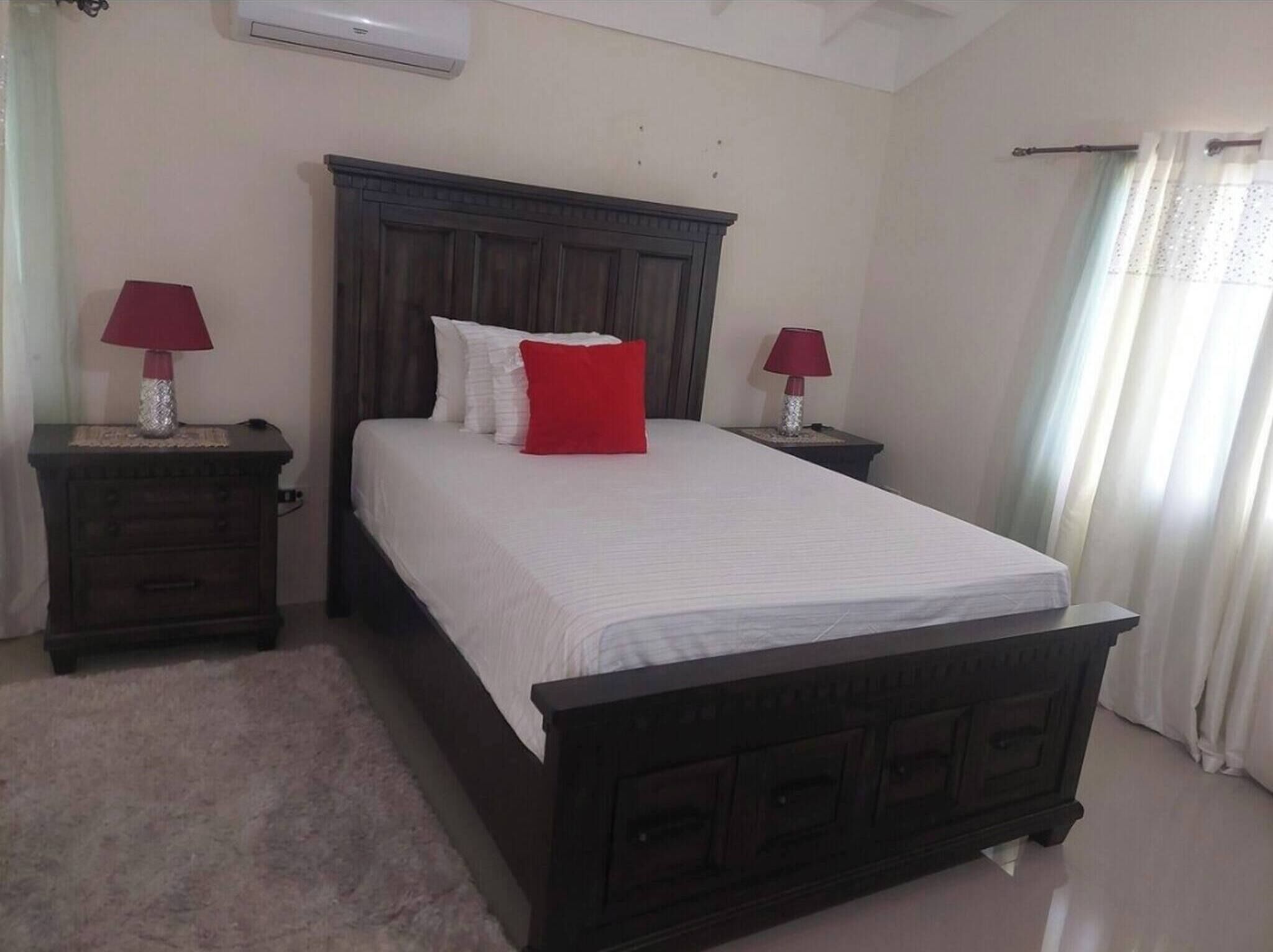 A bedroom with a dark wooden bed, white bedding, two bedside tables with lamps, a red pillow, and a window with curtains.