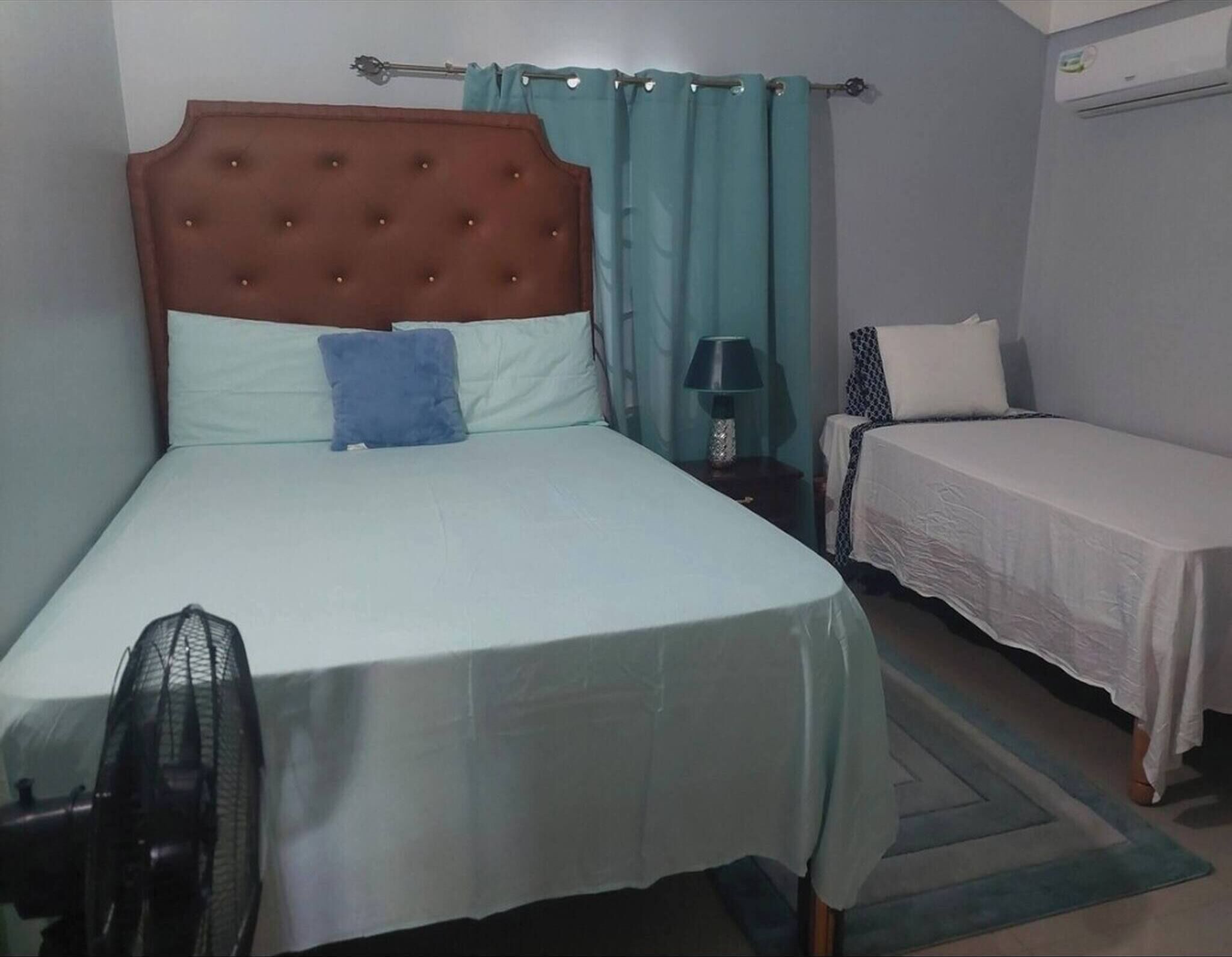 Quarto com cama, mesa de cabeceira, abajur, ventilador e uma pequena mesa com cadeira.
