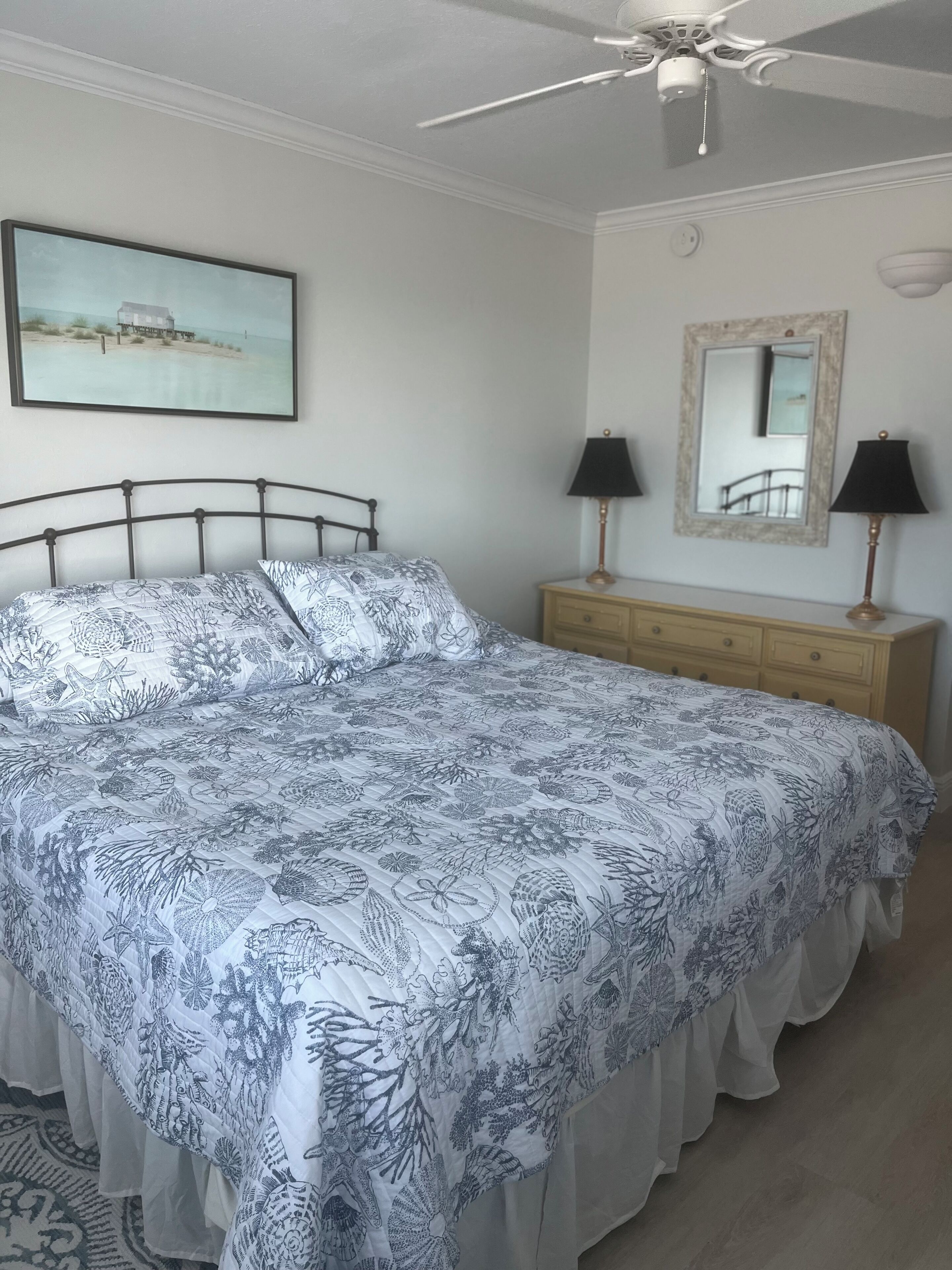 Un dormitorio con una cama, dos lámparas de noche, un aparador, un ventilador de techo, un espejo en marco y un cuadro en la pared.
