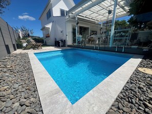 Une piscine rectangulaire bordée de pierres, entourée d’une terrasse aménagée avec des chaises et une table.