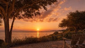 Vista del atardecer sobre una masa de agua, con una silla de madera y un árbol en primer plano.