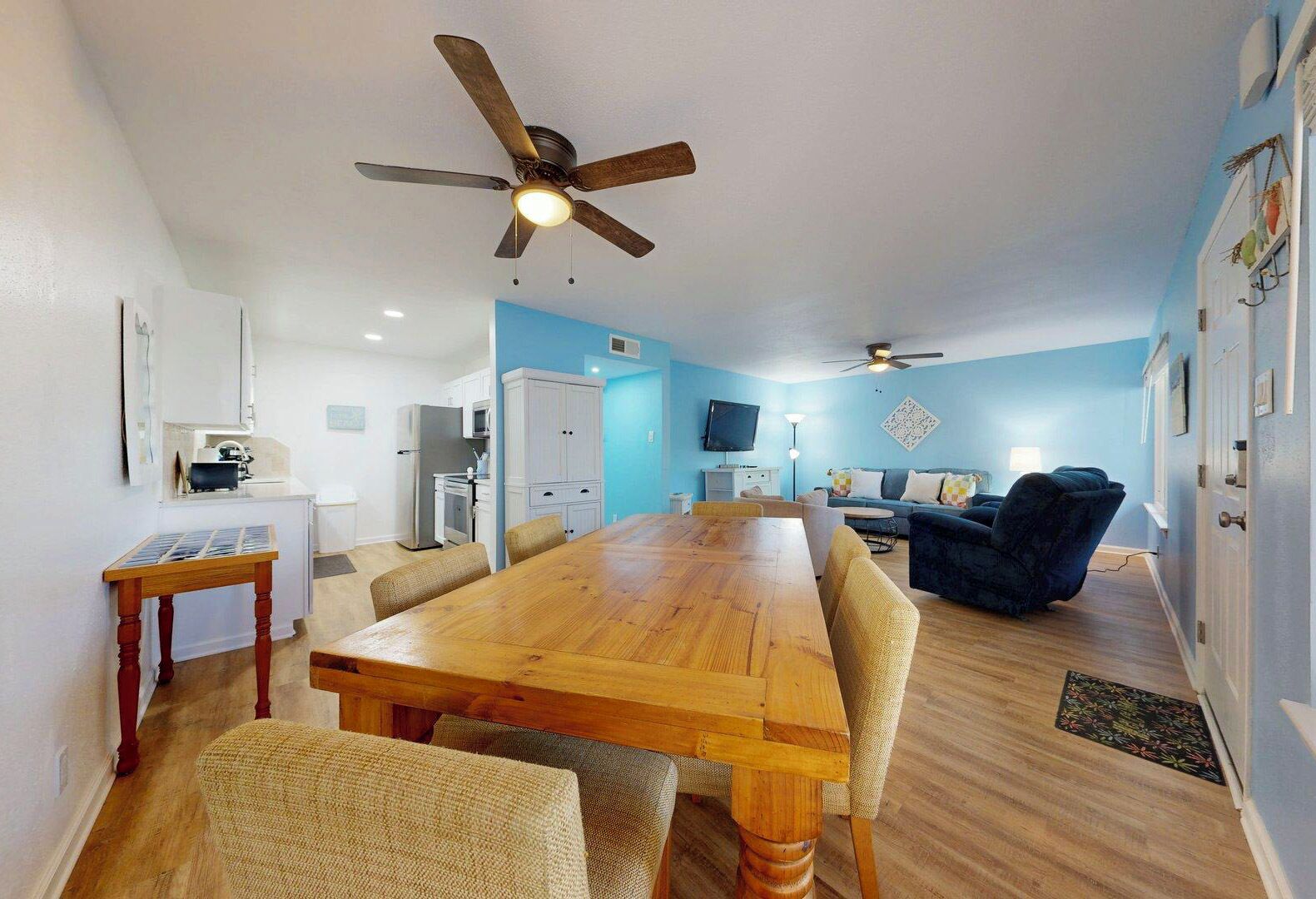 A spacious living area with a wooden dining table, comfortable seating, a ceiling fan, and a kitchen area in the background.