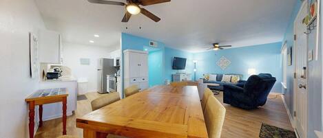 A spacious living area with a wooden dining table, comfortable seating, a ceiling fan, and a kitchen area in the background.