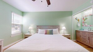 Ein Schlafzimmer mit einem großen Bett, zwei Nachttischen inklusive Lampen, einem Holz-Kopfteil, einem Fenster mit Jalousien und einem Badezimmer mit Waschtisch und Spiegel.