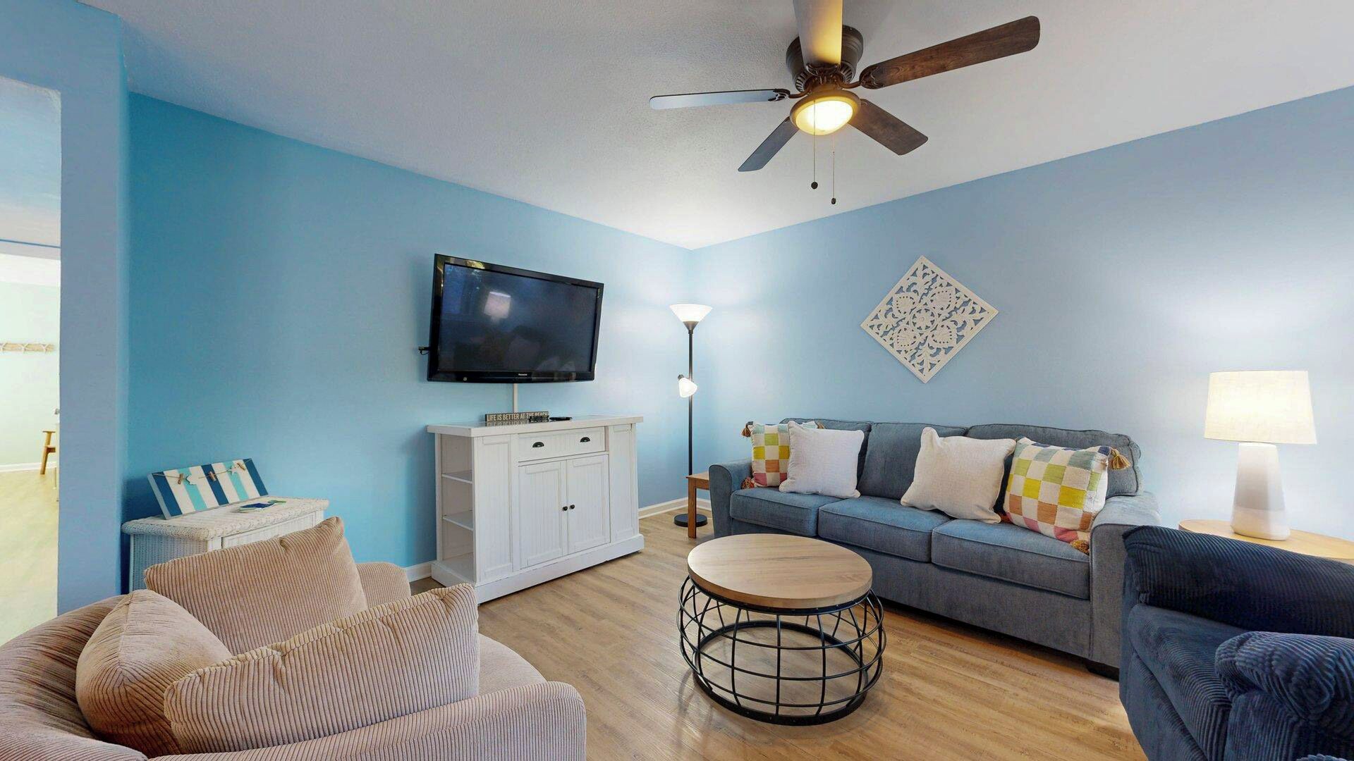 Ein Wohnzimmer mit blauer Wand, Deckenventilator, Flachbildfernseher, einer Couch mit Kissen, einem runden Couchtisch und einem Beistelltisch mit Lampe.