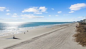 A wide beach with a clear sky, a few people walking, and buildings along the shore. - The High Tide Hideaway @ The Beach  (North Myrtle Beach)