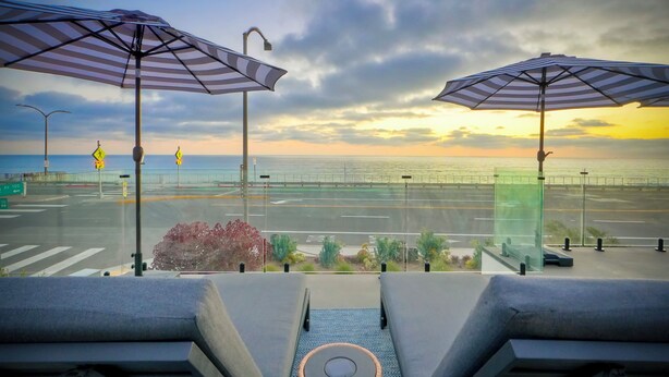 Une vue depuis un balcon, avec deux parasols à rayures, une route et un coucher de soleil sur l’océan.