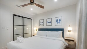 Un lit bien fait, avec du linge de lit blanc, une tête de lit bleue et un ventilateur de plafond. Trois tableaux encadrés ornent le mur, ainsi qu’une table de chevet avec une lampe et une porte-fenêtre coulissante.
