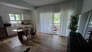 A modern kitchen with white cabinets, a dining area with a wooden table and chairs, and a large window with curtains. - Tracadero Lodge H2 Apt, 1 Bedroom (Dominicus)