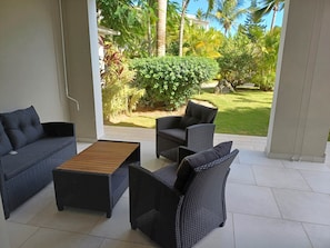 A covered patio area with wicker furniture, a wooden coffee table, and a view of a well-maintained garden with various plants and a lawn. - Tracadero Marina 70 Apt, 1 Hab. (Dominicus)