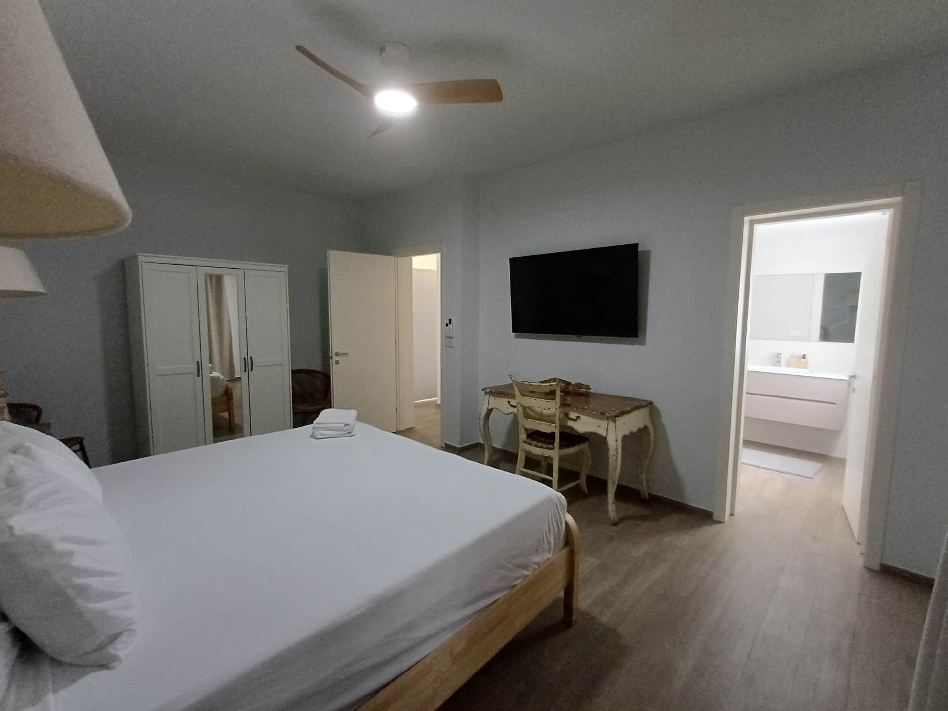 Un dormitorio con cama, escritorio, armario, televisor y baño visible a través de una puerta abierta.