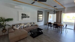 A spacious living room with a sofa, coffee table, and dining area with chairs and a table. A ceiling fan is visible, and there is a window providing natural light. - Tracadero Lodge G3 Apt, 2 Bedroom (Dominicus)