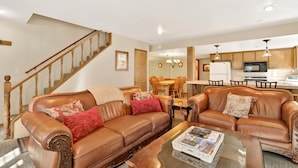 A spacious living room with a brown leather sofa, a coffee table, and a dining area with a wooden table and chairs. A staircase with wooden railings is visible in the background. - Summit Ski Resort Mountain View Condo - Walk to Snow Summit (Big Bear Lake)