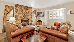 A living room with a stone fireplace, leather sofas, a glass coffee table, and a television mounted on the wall. - Summit Ski Resort Mountain View Condo - Walk to Snow Summit (Big Bear Lake)