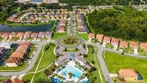 An aerial view of a residential area with a central roundabout, a swimming pool, and houses with red roofs.