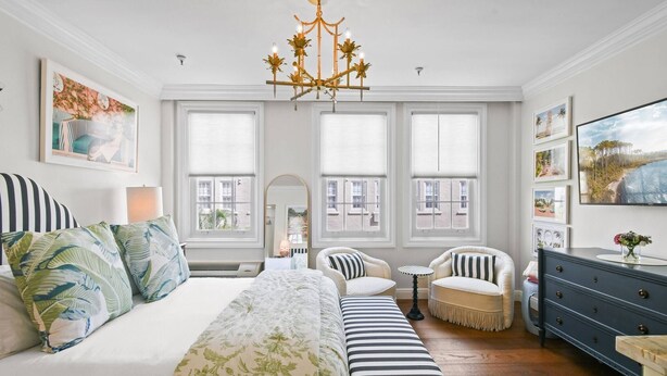 A spacious bedroom with a large bed, a bench, a chair, a dresser, and a chandelier. - Condo Studio/Pool/StepsTO Beach/DINE (Palm Beach)