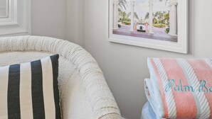 A room with a white wall, a window, a white sofa with striped pillows, and a blue stool with a towel. - Condo Studio/Pool/StepsTO Beach/DINE (Palm Beach)