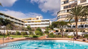Piscina com espreguiçadeiras, rodeada por vegetação e um edifício com varandas.