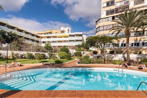 Piscina con tumbonas, rodeada de vegetación y un edificio de varios pisos con balcones.