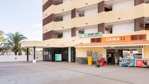 Um edifício de vários andares com área de entrada coberta, um letreiro para 'Holiday Car S.L.' e uma loja 'GAMA' com diversos artigos expostos.