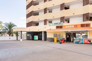 Un edificio de varios pisos con una zona de entrada techada, un cartel que dice 'Holiday Car S.L.' y una tienda 'GAMA' con varios artículos exhibidos en el exterior.