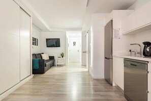 A modern hotel room with a sofa, a small table, a TV, a kitchen area with a dishwasher, and a hallway leading to another room.