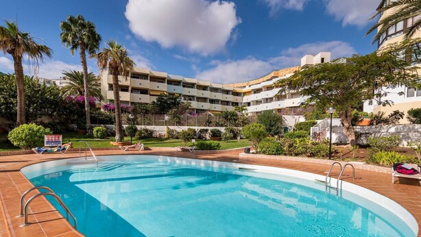 Uma piscina com um deck ladrilhado, rodeada por palmeiras e um prédio de vários andares com varandas.
