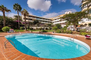 A swimming pool with a tiled deck, surrounded by palm trees and a multi-story building with balconies.