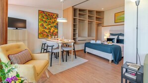Una moderna camera d'albergo con zona pranzo, un letto con copriletto blu, televisione e un grande quadro astratto appeso alla parete.