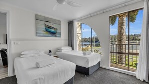 Camera d'albergo con due letti, una ventola a soffitto, un quadro raffigurante una barca e un'ampia finestra che si affaccia su palme e un corpo d'acqua.