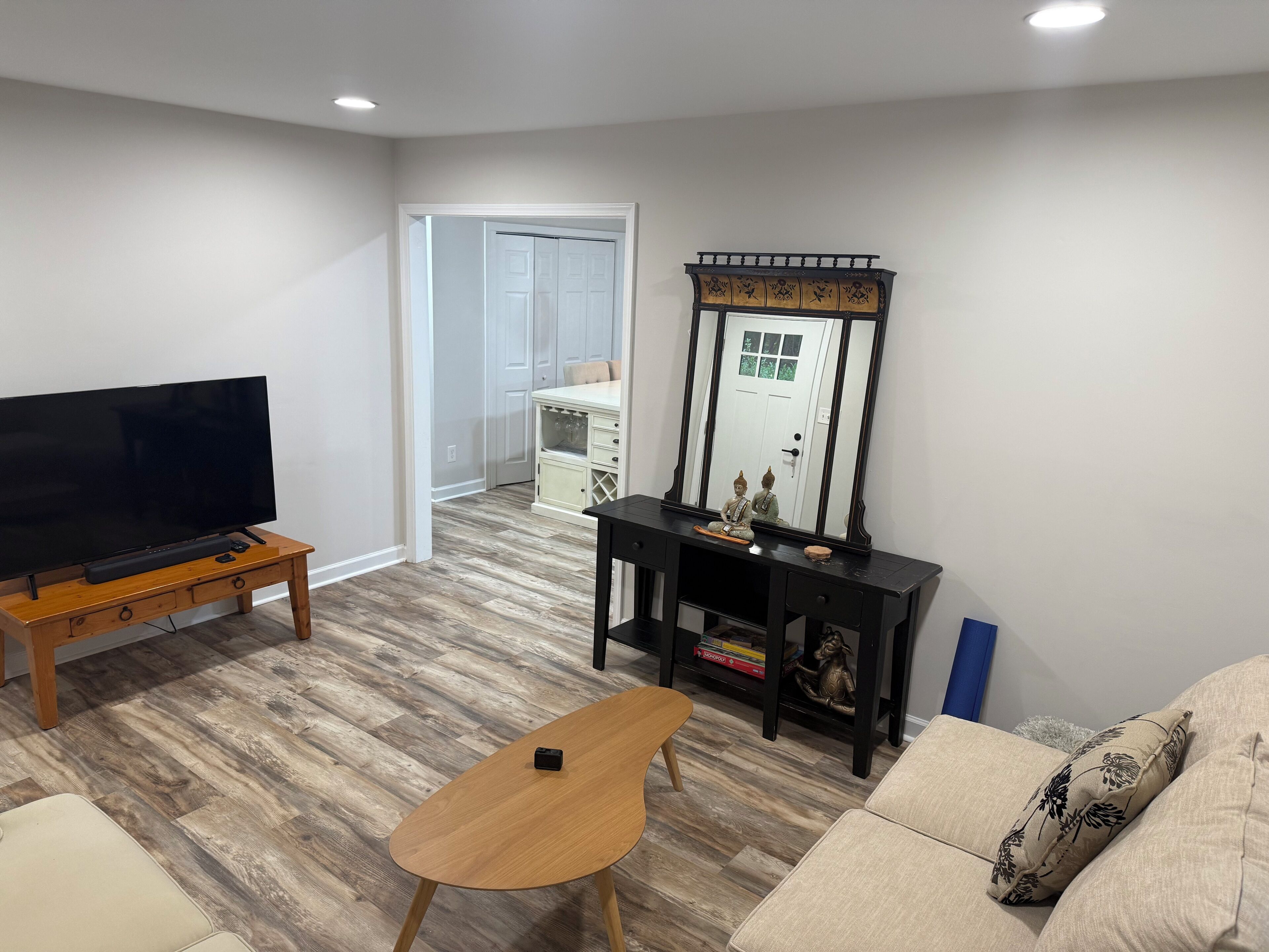 Una sala de estar con un televisor de pantalla plana sobre un pedestal de madera, una mesita de madera pequeña, un sofá con cojines estampados, una consola decorada con objetos decorativos y un espejo grande.