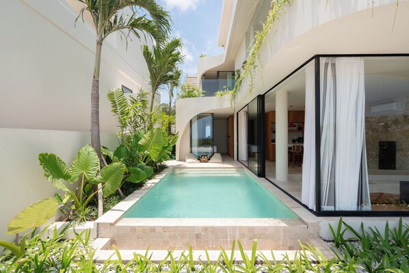 Uma área de piscina moderna com piscina azul límpida, rodeada por exuberantes plantas tropicais e um edifício curvo com grandes janelas de vidro.