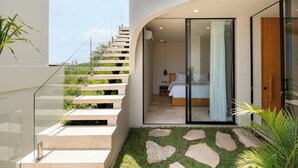 Habitación de hotel moderna con puerta de vidrio que da a un balcón, una escalera con plantas y un pequeño jardín con piedras a modo de camino.