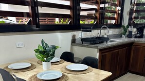 Uma área de refeições com uma mesa de madeira posta para quatro pessoas, cercada por cadeiras pretas. Há uma planta em vasos sobre a mesa, e o cômodo tem vista através de janelas amplas.