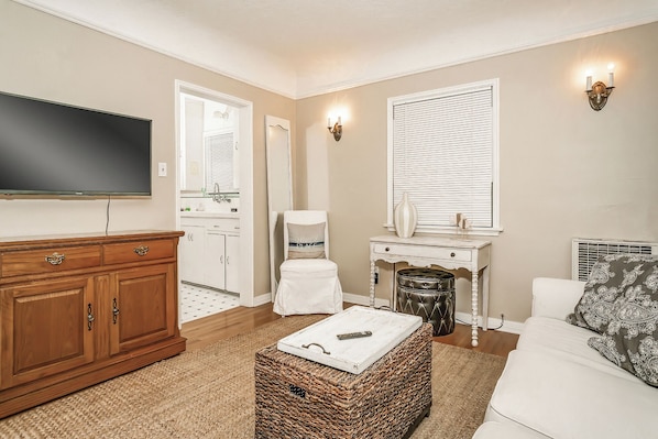 A living room with a television, a wooden cabinet, a wicker ottoman, a white sofa, and a window with blinds.