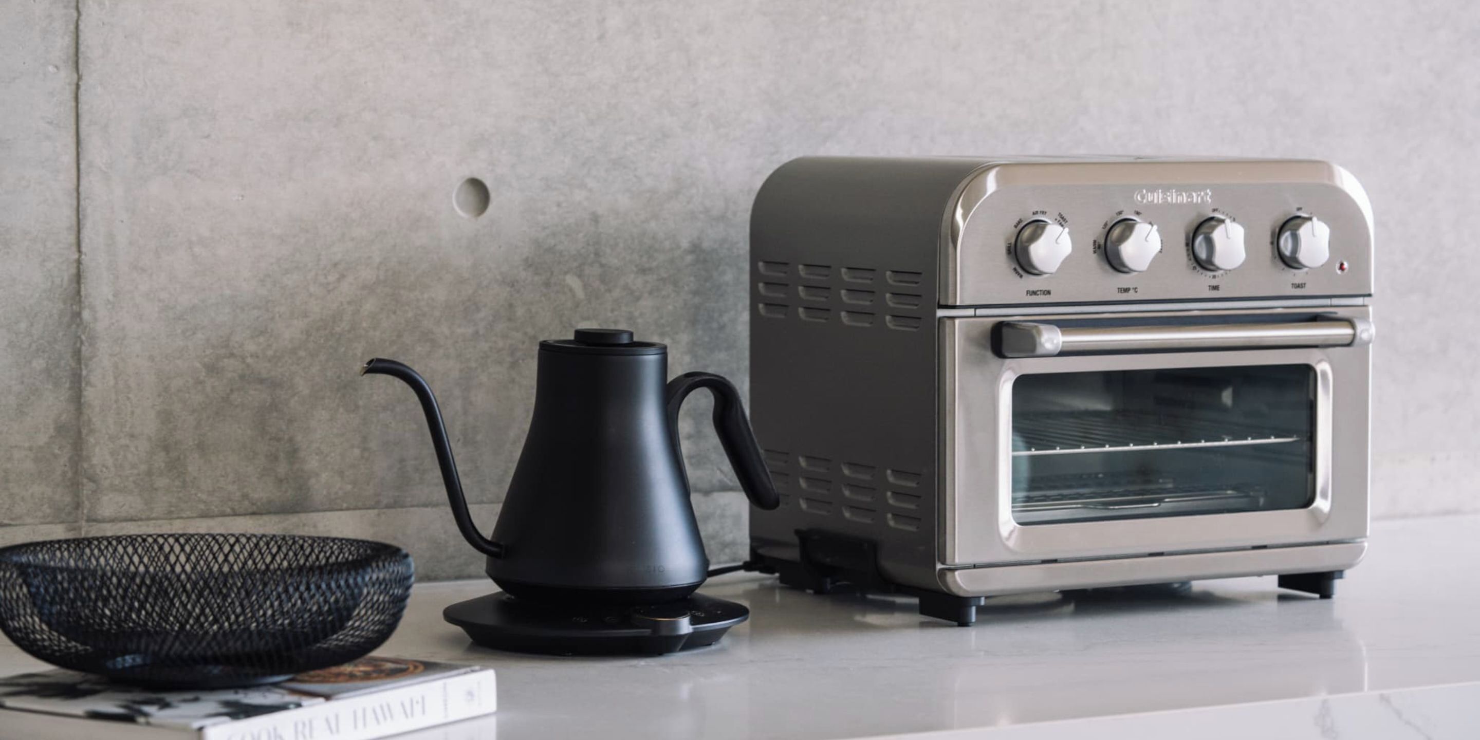A compact kitchen setup with a modern toaster oven, a black electric kettle, and a woven basket on a countertop.