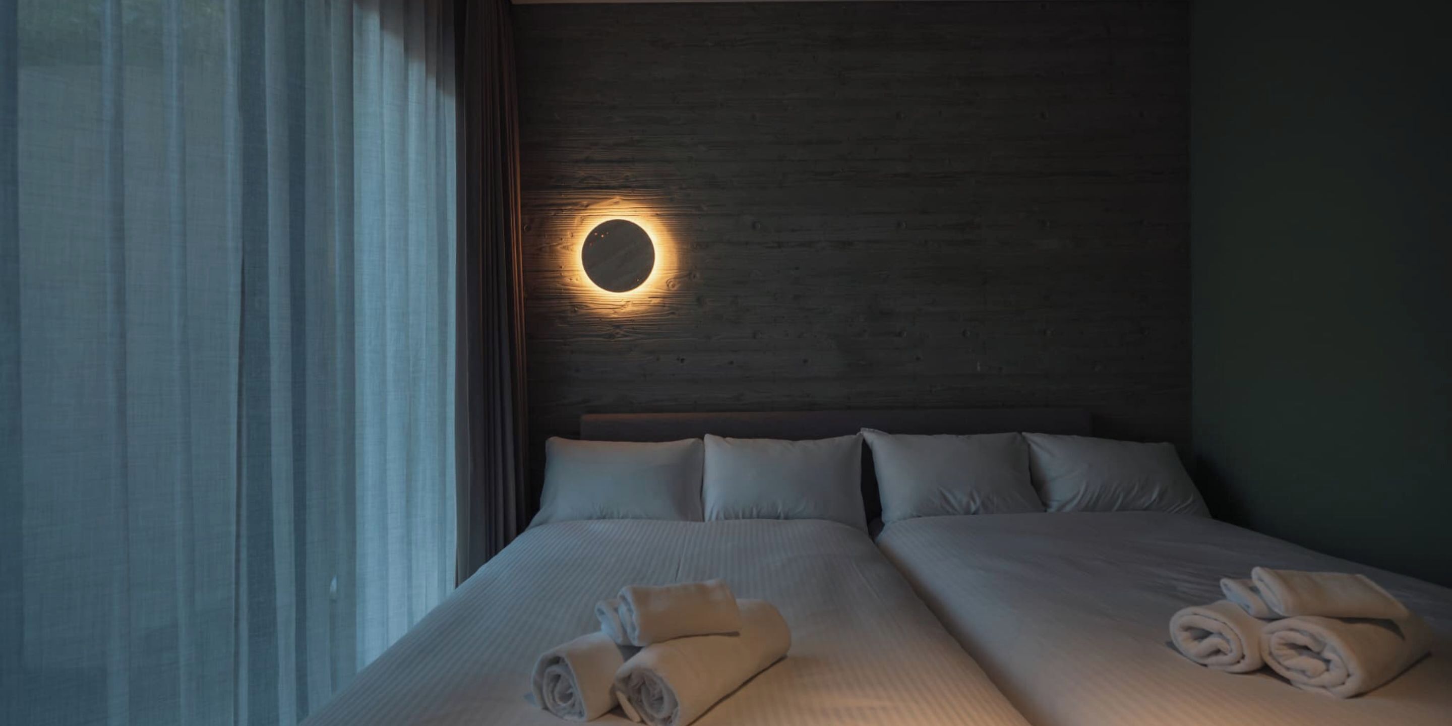 Quarto de hotel com uma cama grande, arrumada com toalhas, uma luminária circular na parede e um painel de cabeceira de madeira.