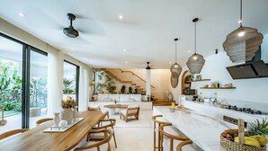 A modern kitchen with a central island, wooden chairs, and a dining area with a large window overlooking greenery. - Kayana Villa Uluwatu (Pecatu)