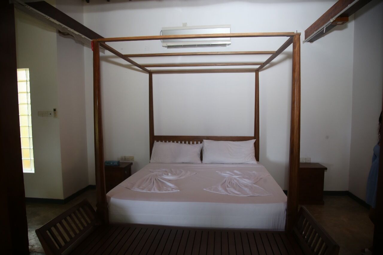 Een hemelbed met een houten frame en een witte matras. Er staat een nachtkastje met een lamp en een klein tafeltje naast het bed.