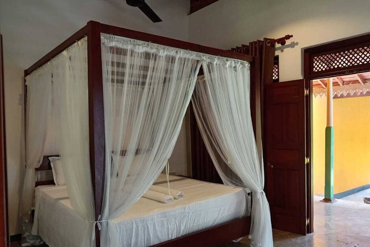 Una cama con dosel de madera, cubierta con una colcha blanca y cortinas traslúcidas, junto a una mesita de noche con una lámpara.