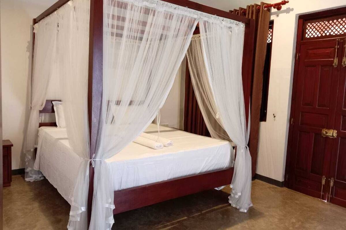 Una cama con dosel con ropa de cama blanca y cortinas translúcidas, suelo de madera y una puerta roja al fondo.