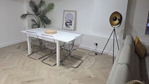 Sala de estar moderna com sofá branco, mesa de jantar com cadeiras transparentes, uma planta em vaso e um abajur.