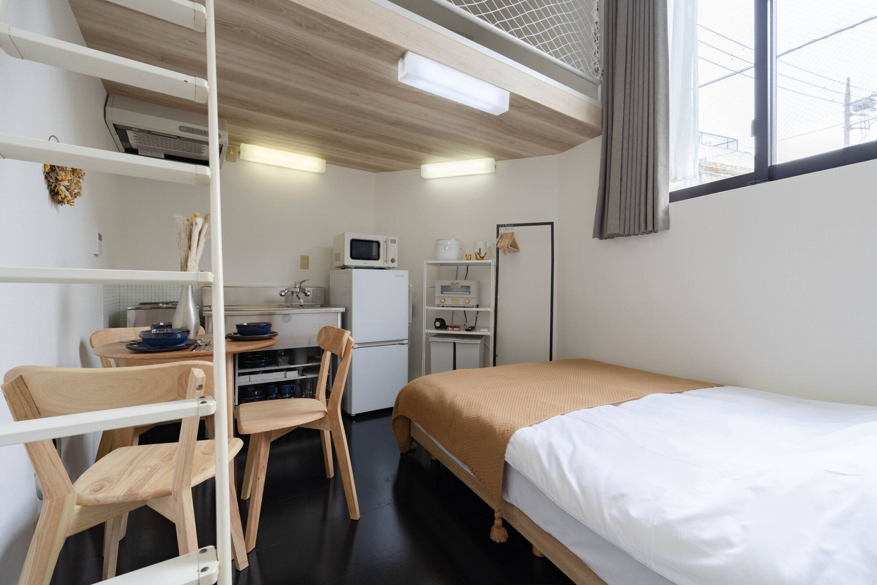 Ein kleines Zimmer mit Etagenbett, einer Küchenzeile, einem Schreibtisch mit Stuhl und einem Fenster mit Vorhängen.