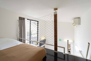 A hotel room with a bed, a window with a metal screen, a wooden column, a wall-mounted light, and a door with an emergency exit sign. - House Hotel nakano TNA (Tokyo)