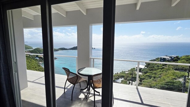 A balcony with a table and chairs, overlooking a coastal view with greenery and a clear sky.