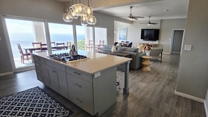 A modern kitchen with a central island, a dining area with a view, and a living room with a television and artwork.