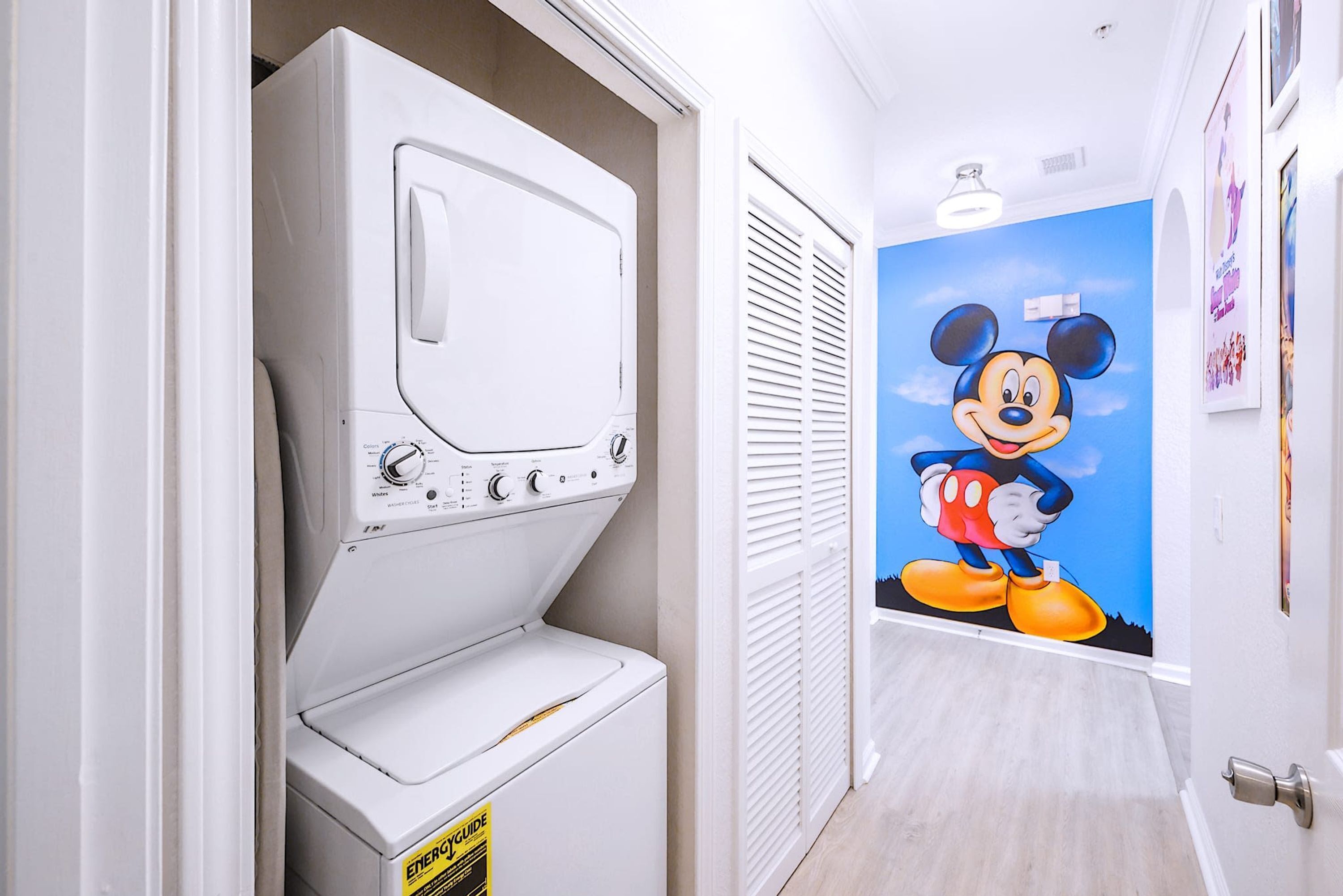 Un lavadero con una lavadora y secadora grandes de color blanco, un mural de Mickey Mouse en la pared y una puerta con manija.